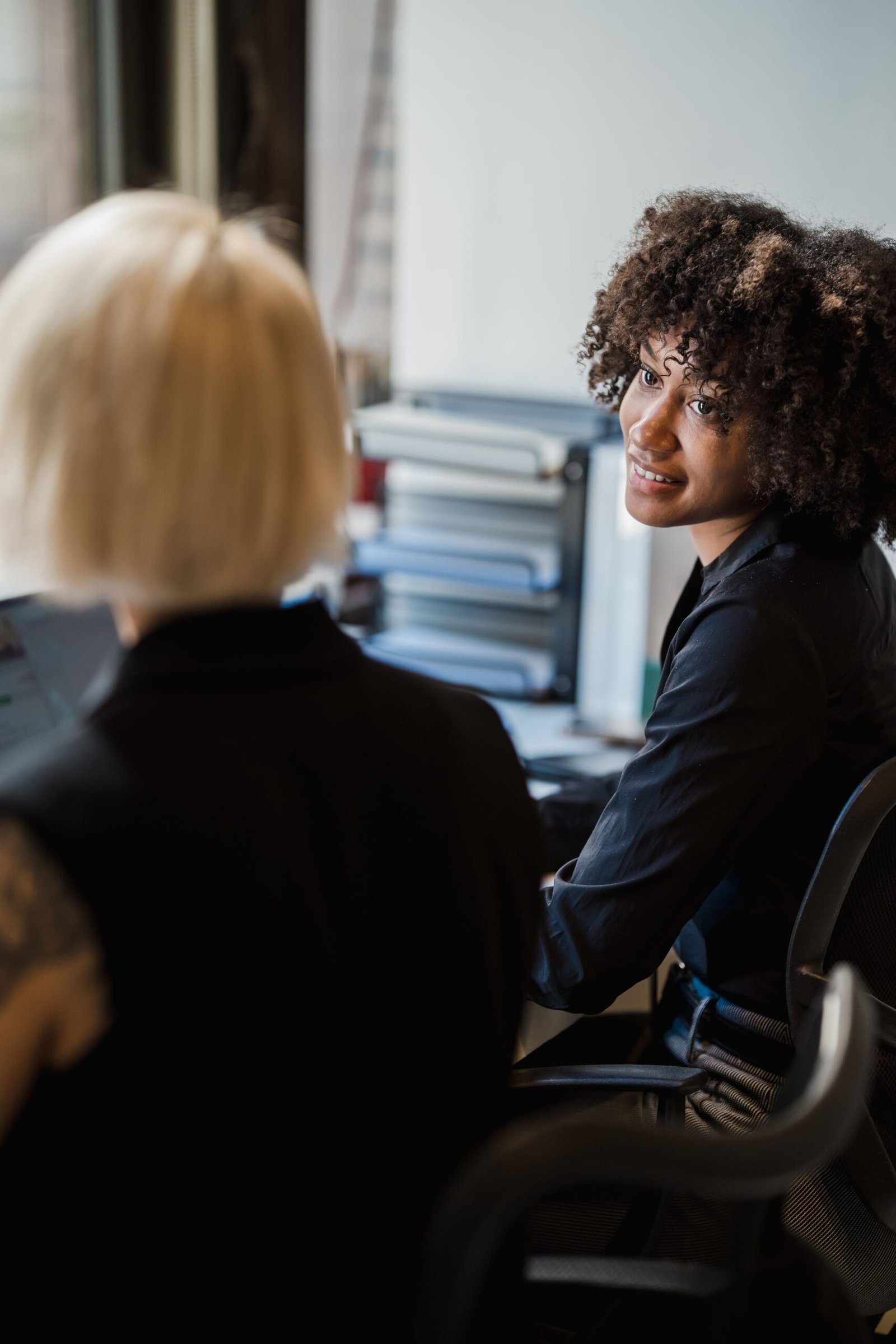 Two professional women engaging in conversation at an office desk, fostering teamwork.